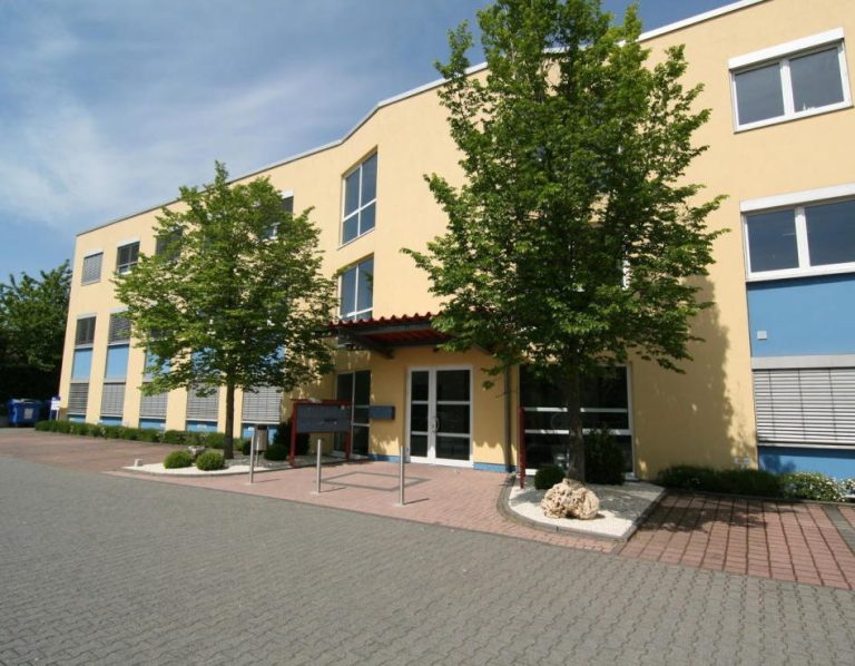 COMA PACPAGUNG GmbH Gelbes Gebäude mit großen Fenstern, umgeben von Bäumen und einem gepflasterten Gehweg.